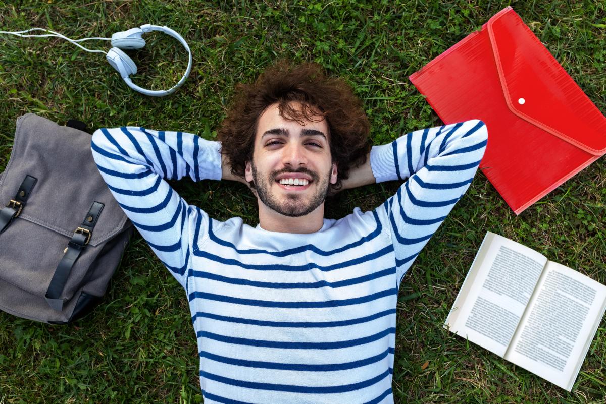 Junger Mann liegt lachend und in den Himmel schauend auf grünem Rasen, neben sich ein geöffnetes Buch, Kopfhörer, Rucksack und Dokumententasche.