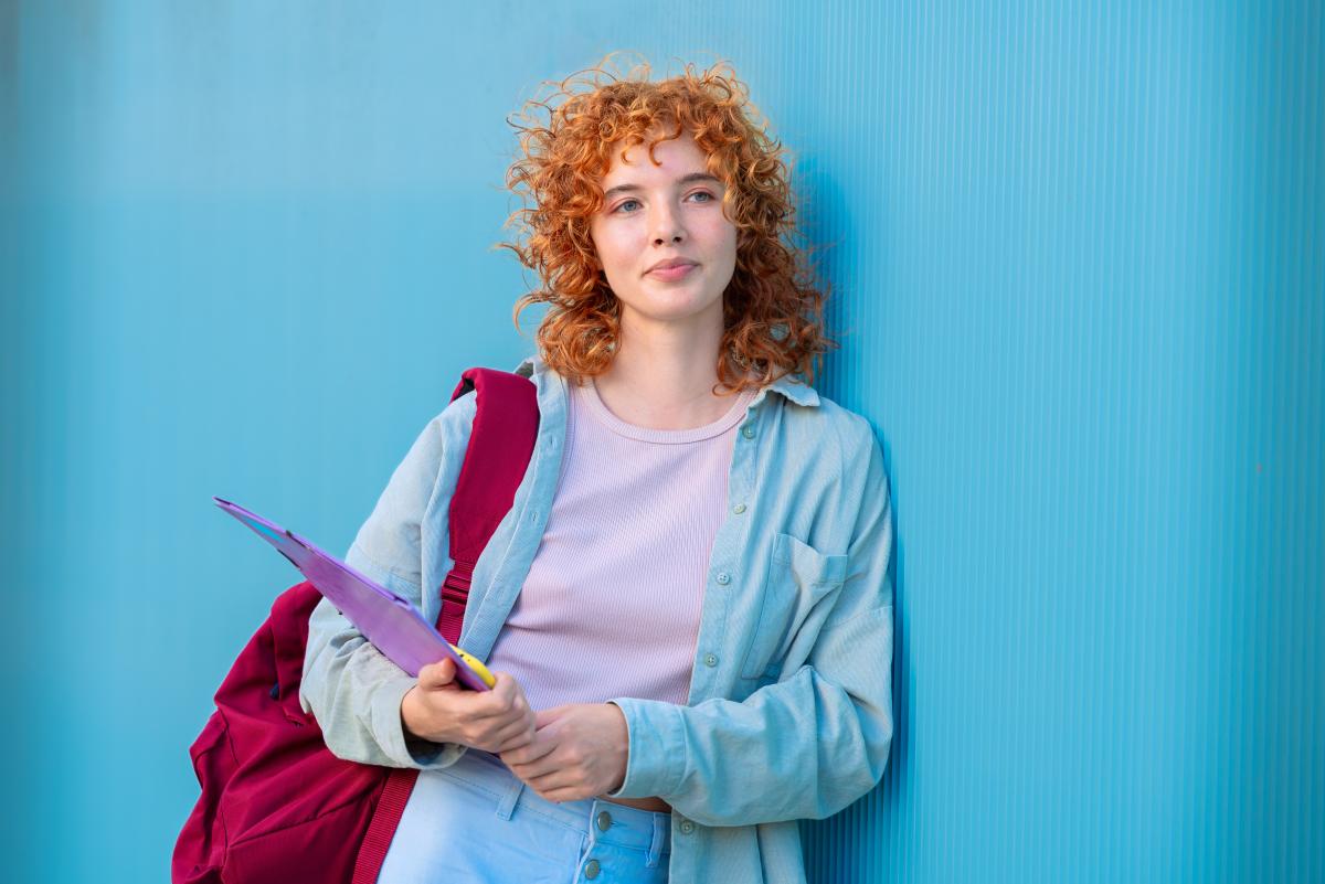 Junge Frau mit Rucksack und Dokumentenhülle lehnt an einer blauen Wand.