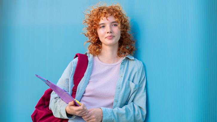 Junge Frau mit Rucksack und Dokumentenhülle lehnt an einer blauen Wand.
