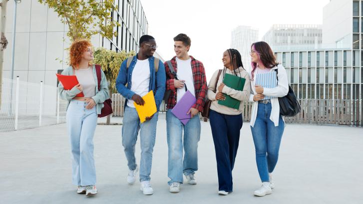 Fünf Studierende mit Heftern spazieren lachen und sich unterhaltend über den Campus.