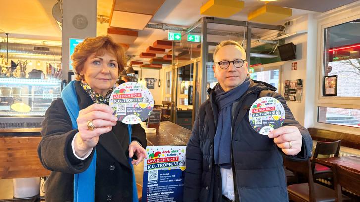 Die Opferschutzbeauftragte Barbara Havliza und Minister Dr. Limbach zeigen Bierdeckel mit Informationen zu K.O.-Tropfen Missbrauch